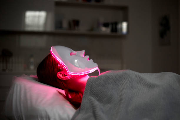 A person lies on a bed wearing a glowing red light therapy LED mask for skincare benefits.